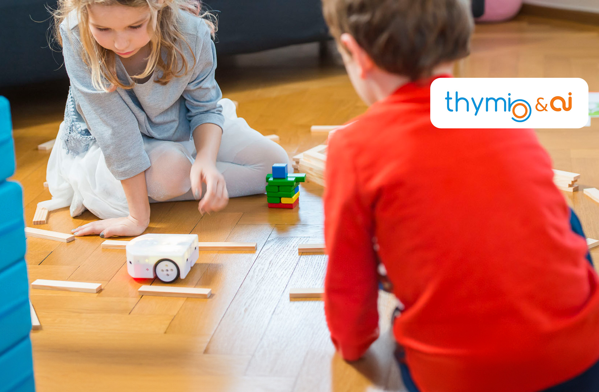 LP Header Thymio Zwei Kinder spielen mit dem Thymio Lernroboter auf dem Boden und lassen diesen durch einen Parcours fahren. Oben rechts ist das Thymio Logo zu sehen.