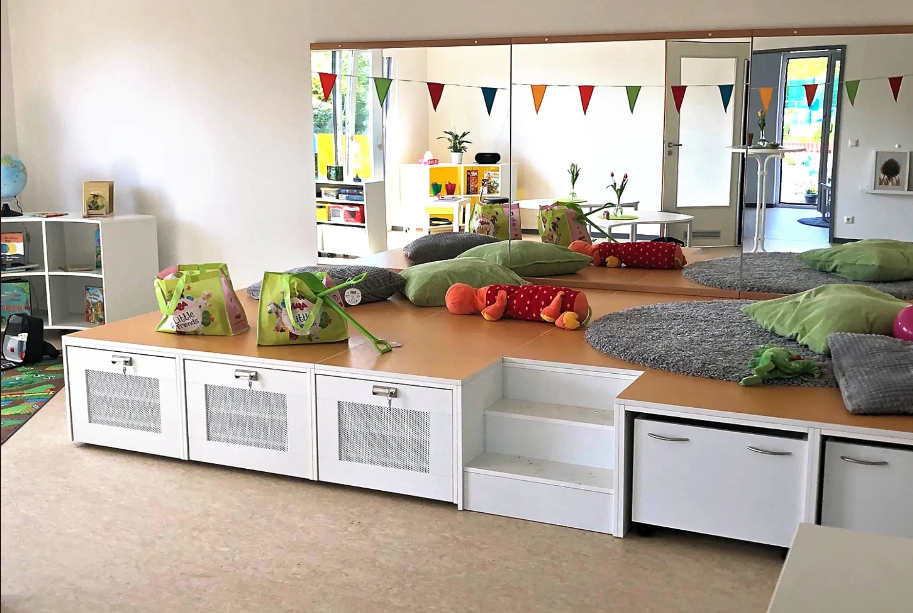 Marlower Möbel Kindergarteneinrichtung Auf dem Bild ist eine Kuschelecke in einem Kindergarten zusehen. Ein kleines Podest ist aufgebaut, sodass sich die Kuschelecke etwas höher als der Boden befindet. Auf dem Podest sind mehrere grüne und graue Kissen, ein rotes Kuscheltier und ein grauer Tep